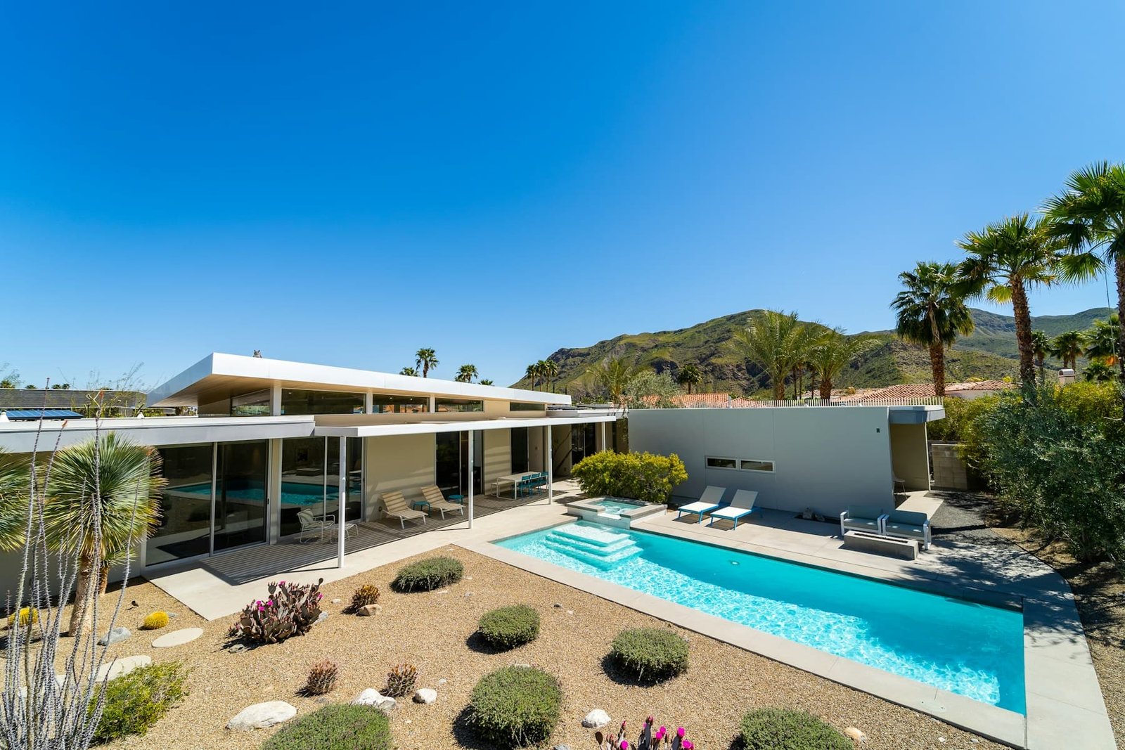 A mid-century modern home with a rectangular pool, raised spa, palms and mountains behind it under a clear Southern California sky