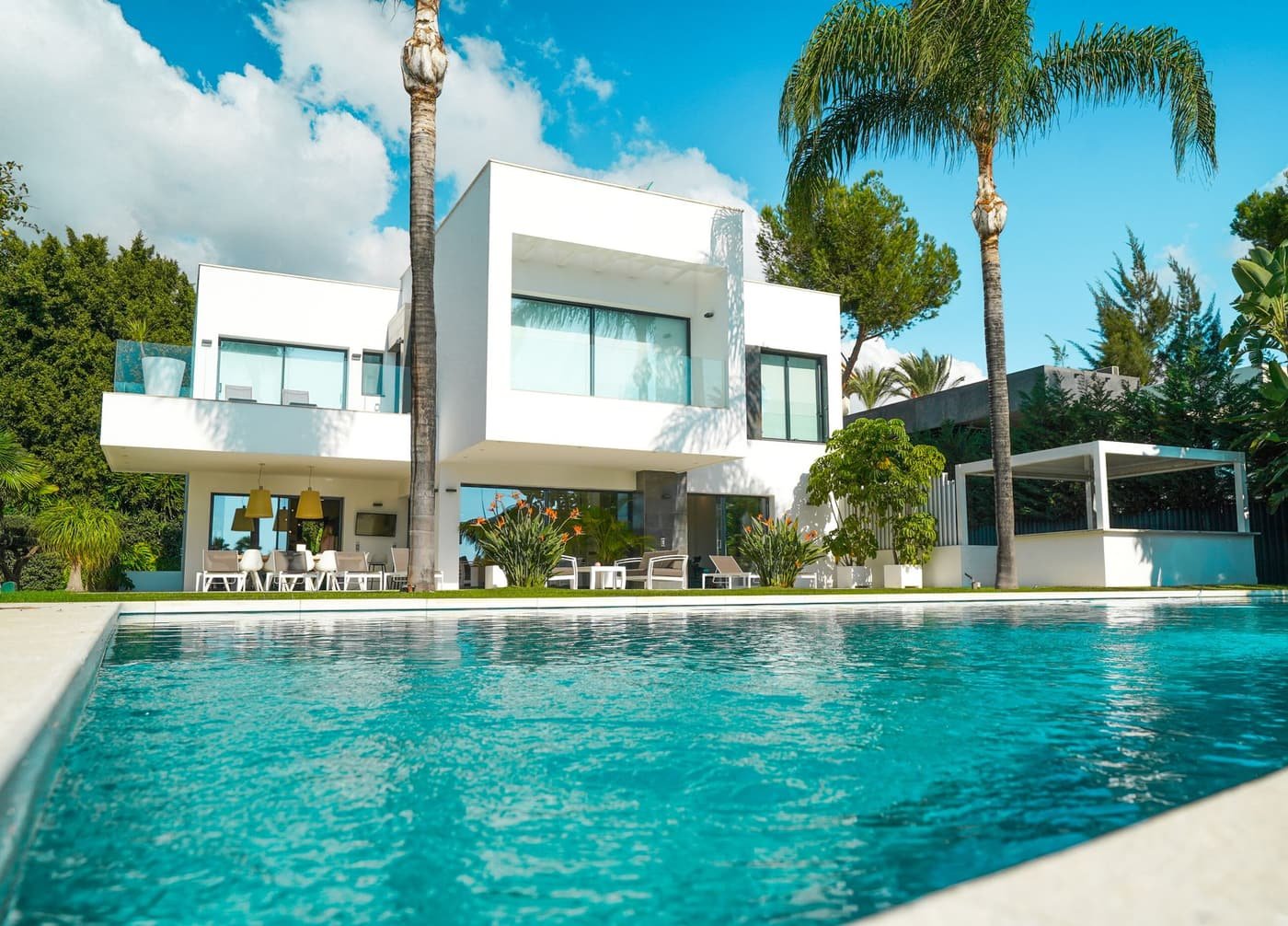 A long rectangular pool with a fresh finish in front of a white modern home and palms